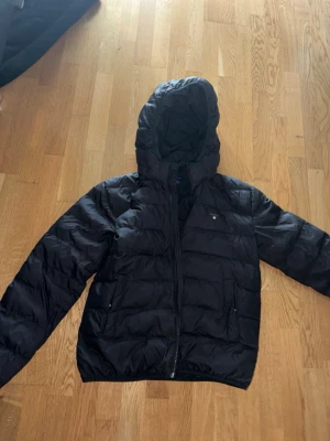 Svart dunjacka med huva från GANT - Äkta Snygg svart dunjacka från GANT med huva och dragkedja framtill köpt från kidsbrandstore. Jackan har två sidofickor och diskret GANT-logga på bröstet. Perfekt för kalla vinterdagar och har en klassisk, puffig design.
