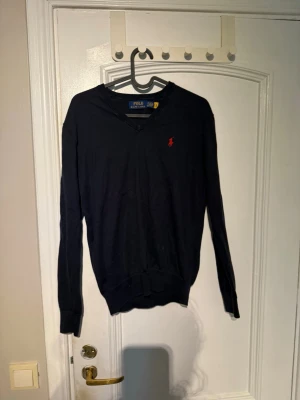 Mörkblå V-ringad tröja Polo Ralph Lauren - Mörkblå V-ringad tröja från Polo Ralph Lauren med den klassiska röda logotypen broderad på bröstet. Tröjan har långa ärmar och ribbade muddar vid ärmslut och nederkant. Perfekt för en stilren och enkel look.