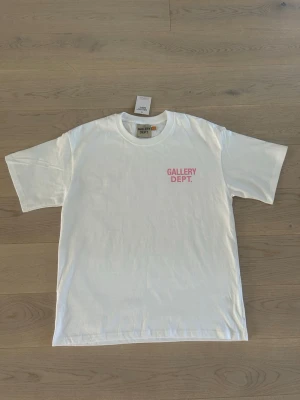Vit Gallery Dept. t-shirt med rosa tryck - Vit t-shirt från Gallery Dept. med rosa texttryck fram och stort tryck på ryggen där det står 'GALLERY DEPT. HOLLYWOOD, CA.'. Klassisk passform och rund hals. Tillverkad i mjuk bomull, perfekt för en avslappnad stil. Är i storlek M men sitter som storlek L!
