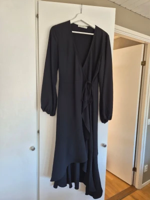 Carin Wester lång omlottklänning stl 38 - Snygg svart långklänning med omlottmodell och knytband i midjan. Klänningen har långa ärmar med lätt puff och v-ringad hals. Tyget faller fint och ger en elegant siluett. Perfekt för dig som gillar stilrena och tidlösa plagg. Nyskick ✨️✨️