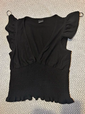 Svart volangblus från Gina Tricot - Snygg svart blus från Gina Tricot med v-ringning, volangdetaljer vid ärmslut och nederkant samt smockad midja. Toppen har korta ärmar och är tillverkad i ett mjukt, lätt material som ger en skön passform.