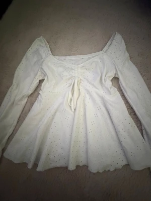 Vit blus med puffärm och knyt - Supersöt vit blus med långa puffärmar och smockad detalj framtill med knytband. Blusen har små hålmönster över hela tyget och en lätt utsvängd passform. Perfekt för dig som gillar romantisk och trendig stil.