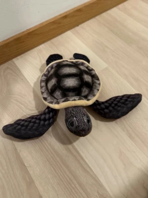 Mjukisdjur sköldpadda realistisk - Säljer en mjukisdjurssköldpadda med realistiskt utseende. Sköldpaddan är svart, grå och beige med detaljerad sköld och fenor. Tillverkad i mjukt tyg och stoppad med fluffigt material. Perfekt som prydnad eller gosedjur. Aldrig använd, vunnen på Gröna Lund. 