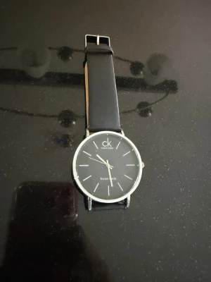 Svart klocka från Calvin Klein - Stilren klocka från Calvin Klein med svart urtavla och silverfärgade detaljer. Klockan har ett tunt, runt boett i silver och ett klassiskt svart läderband. Minimalistisk design med tydliga markeringar och logga på urtavlan.