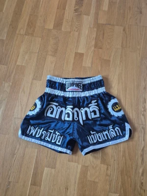 Muay Thai shorts från Lumpinee - Snygga Muay Thai shorts från Lumpinee i blankt svart material med vita och gula detaljer samt thailändsk text. Shortsen har bred resår i midjan och kontrasterande kantband. Perfekta för träning eller match inom kampsport.