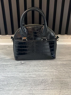 Svart handväska i krokopräglat skinn från Zara - Snygg svart handväska med krokodilpräglat mönster och rundade handtag. Väskan har en stilren, halvmåneformad design och silverfärgade metalldetaljer. Dragkedja upptill och ringar för axelrem. Perfekt för dig som gillar trendiga accessoarer. Den är fin och är från ZARA