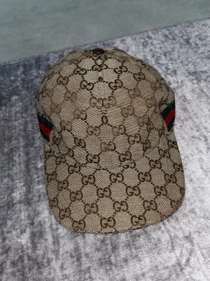 Gucci beige keps med GG-mönster - Säljer en beige keps från Gucci med det klassiska GG-monogrammet över hela kepsen. På sidan finns en grön och röd rand som ger extra lyxig känsla. Kepsen är tillverkad i canvasmaterial och har en böjd skärm. Perfekt för dig som vill sticka ut med en ikonisk accessoar.