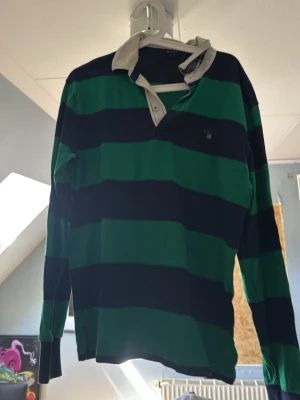 Randig rugbytröja från GANT - Säljer en  äkta klassisk långärmad rugbytröja från GANT med breda gröna och marinblå ränder. Tröjan har vit krage och knappslå vid halsen. Perfekt för en sportig och avslappnad stil. 