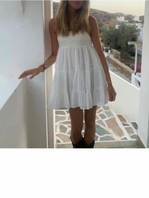 Vit mini sommarklänning - Söt vit kort klänning med smala axelband och smockad överdel. Kjolen är luftig och har volanger för en flowy känsla. Perfekt för varma dagar och enkel att styla med boots eller sandaler.