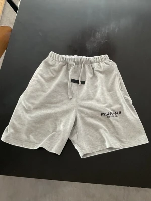 Grå Essentials Fear of God shorts - Säljer ett par ljusgrå shorts från Essentials Fear of God med snörning i midjan och tryckt logga på benet. Shortsen har en avslappnad passform och är tillverkade i mjuk bomull, perfekta för chill dagar.