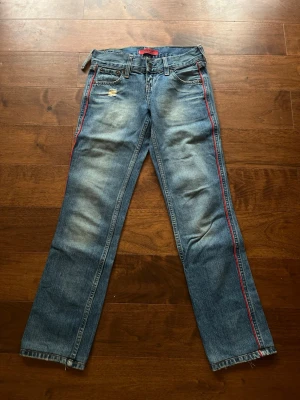 Levi’s jeans  - Snygga blå Levis jeans  med raka ben och en röd rand längs sidorna. Jeansen har slitna detaljer vid fickan och klassisk femficksdesign. En hälla är trasig men går lätt att sy. Jeansen är min mammas gamla och är försmå på mig, därav ingen bild med de på. Mått:25x32
