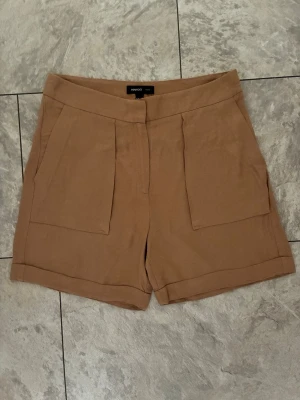 Beige shorts från Mango med fickor storlek 36 - Stilrena beige shorts från Mango med hög midja och stora framfickor. De har dold dragkedja och knappstängning framtill. Perfekta för varma dagar och enkla att matcha med olika toppar.