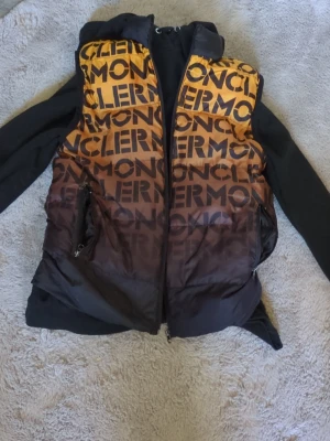 Moncler dunväst med logotypmönster - (kolla 3dje bilden)Snygg dunväst från Moncler i svart och gul-orange gradient med stort logotypmönster över hela västen. Västen har hög krage, två dragkedjeförsedda fickor och hel dragkedja framtill. Perfekt för dig som vill sticka ut med en trendig och exklusiv look.