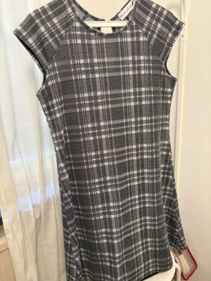 Gråvit rutig kortklänning - Snygg gråvit kortklänning med rutigt mönster och rund hals. Klänningen har korta ärmar och en liten öppning med knapp i nacken. Perfekt för dig som gillar klassisk stil med en modern twist.