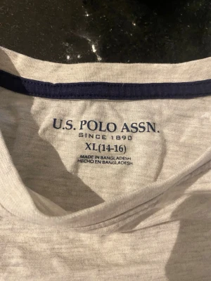 Ljusgrå t-shirt från U.S. Polo Assn. - Säljer en ljusgrå t-shirt från U.S. Polo Assn. med mörkblå broderad logga på bröstfickan och kontrastfärgad kant i halsen. Tillverkad i mjuk bomull, perfekt för en avslappnad och stilren look.