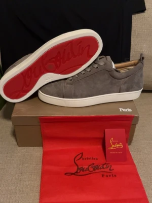 Gråa Christian Louboutin sneakers - Snygga gråa sneakers från Christian Louboutin i mocka med klassisk röd sula och vita kanter. Lågt skaft och diskret design med ton-i-ton snörning. Insidan är beige och skon har exklusiva detaljer som sticker ut.