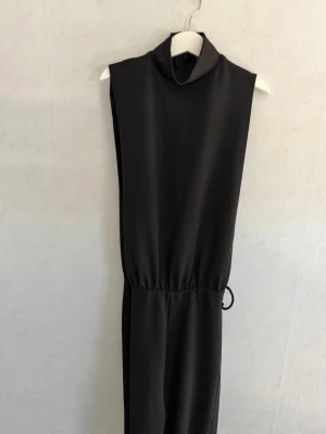 Svart ärmlös jumpsuit med polokrage - Snygg svart ärmlös jumpsuit med hög polokrage och resår i midjan. Tillverkad i ett mjukt, stretchigt material som ger en clean och modern look. Perfekt för dig som gillar minimalistisk stil och vill ha något bekvämt men ändå trendigt.
