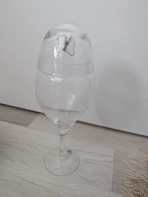 Dekorations glas med fjärilsdekor, ska vara dekor sand eller liknande - Dekorations glas som det har varit dekor sand i innan. I genomskinligt glas med en dekorativ fjäril i lila. Glaset har klassisk form med rund kupa och smal fot. Perfekt för dig som gillar detaljer och vill skapa något dekorativt till hemmet.