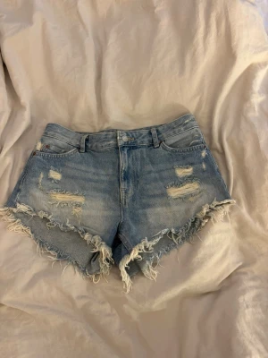 Ljusblå jeansshorts med fransig kant - Säljer ett par ljusblå denimshorts med fransig kant och slitna detaljer framtill. Klassisk femficksmodell med knapp och dragkedja. Perfekta för sommaren och ger en avslappnad vibe.