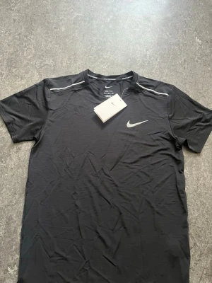 Nike Miller t shirt  - Sportig svart t-shirt från Nike med vit logga på bröstet och tunna vita linjer vid axlarna. Tillverkad i lätt och funktionell polyester som andas, perfekt för träning. Klassisk rund hals och korta ärmar för en clean och modern look.