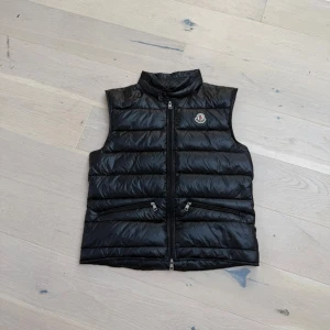 Svart dunväst från Moncler - Snygg svart dunväst från Moncler med hög krage och två dragkedjefickor framtill. Västen har ett quiltat mönster och ikonisk Moncler-logga på bröstet. Perfekt för lager-på-lager och ger en clean look till din outfit.