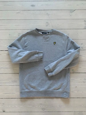 Grå sweatshirt från Lyle & Scott - Klassisk grå sweatshirt från Lyle & Scott med rund halsringning och gul logga på bröstet. Tröjan har ribbade muddar vid ärmslut och nederkant, samt en relaxed passform. Perfekt för en avslappnad och stilren look.