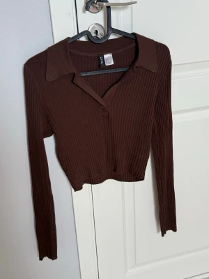 Brun ribbad crop top från H&M - Säljer en brun ribbad långärmad crop top från H&M. Tröjan har krage, v-ringning och är tillverkad i ett stretchigt material som sitter snyggt på kroppen. Perfekt till jeans eller kjol för en trendig look.