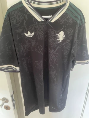 Svart Adidas fotbollströja Ezhegrova - Snygg svart fotbollströja Juventus från Adidas med diskret bladmönster, vita detaljer och randiga muddar. Tröjan har tryck med namn och nummer 'E.ZHEGROVA 11' på ryggen samt Adidas-logga och klubbmärke på bröstet. Tillverkad i lätt och ventilerande material. 