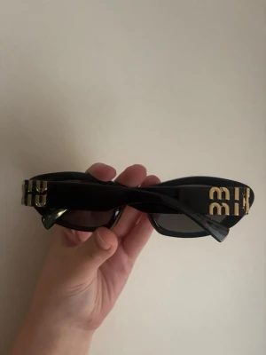 Miu Miu solglasögon - Snygga svarta solglasögon från Miu Miu med breda bågar och guldiga BB-detaljer på sidorna. Glasen är mörka och bågarna har en oval, lite cat-eye form som ger en cool vibe. 