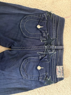 Mörkblå True Religion jeans - Säljer ett par mörkblå jeans från True Religion med ikoniska kontrastsömmar och stora bakfickor med dekorativa knappar. Jeansen har bootcut-modell och är tillverkade i denim med coola detaljer på fickorna. Perfekta för dig som gillar streetwear och vill sticka ut. Säljer pga att de är för stora.