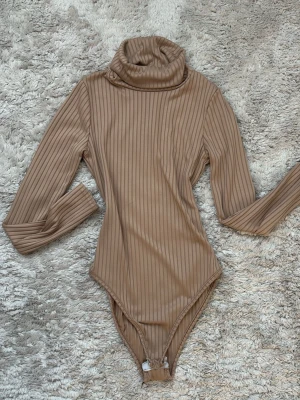 Beige ribbad body från Boohoo - Snygg beige body från Boohoo med ribbad struktur och polokrage. Långärmad modell i mjukt stretchigt material som sitter skönt mot kroppen. Perfekt att styla med jeans eller kjol, otroligt elegant! Gåva vid köp!!