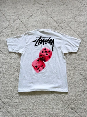 Vit Stüssy t-shirt med tärningsmotiv - Vit t-shirt från Stüssy med stort tryck på ryggen som visar två rosa tärningar och Stüssy-loggan i svart. Klassisk passform och korta ärmar. Perfekt för dig som gillar streetwear och vill sticka ut med en unik design.
