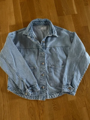 Ljusblå jeansjacka från Zara - Klassisk ljusblå jeansjacka från Zara med raka linjer, två stora framfickor och silverfärgade knappar. Jackan har en avslappnad passform och en snygg krage. Perfekt att slänga över en hoodie eller t-shirt för en cool och enkel look.