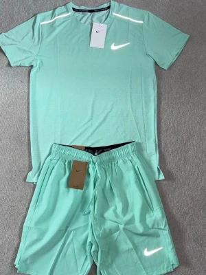 Mintgrönt träningsset från Nike - Snyggt mintgrönt träningsset från Nike med t-shirt och shorts. Båda plaggen har reflekterande Nike-loggor och är tillverkade i ett lätt, snabbtorkande material som passar perfekt för löpning eller gym. T-shirten har rund hals och korta ärmar, shortsen har resår i midjan.