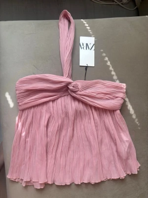 Rosa omlott topp från Zara - Supersöt rosa omlott topp från Zara med plisserat tyg och bred axelrem. Toppen har en knutdetalj framtill och är tillverkad i ett lätt, luftigt material som ger en flowy känsla. Perfekt för dig som vill ha en trendig och feminin look.