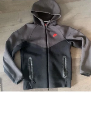 Mörkgrå Nike hoodie med dragkedja - Säljer en snygg mörkgrå hoodie från Nike med hel dragkedja och huva. Tröjan har två fickor med dragkedja framtill och en röd Nike-logga på bröstet. Tillverkad i mjukt material som passar perfekt för en avslappnad streetwear-look.
