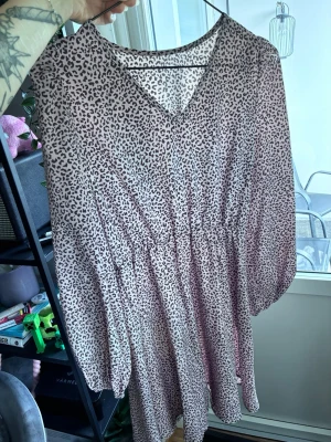 Leopardmönstrad kortklänning - Säljer en snygg kortklänning med leopardmönster i ljusrosa och svart. Klänningen har v-ringning, långa ärmar och resår i midjan för en skön passform. Perfekt för dig som vill sticka ut lite extra!