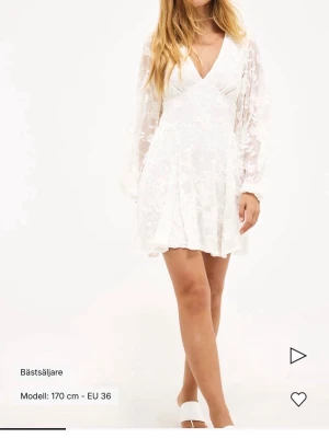 Vit blommig kortklänning med öppen rygg - Supersöt vit kortklänning med blommigt mönster, v-ringning och transparenta långärmar. Klänningen har öppen rygg med knytband och volangkant nertill. Perfekt för sommarens alla tillfällen!