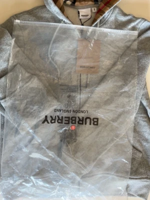 Grå hoodie med dragkedja från Burberry - Snygg grå hoodie från Burberry med dragkedja och klassiskt rutigt foder i huvan. Broderad logga på bröstet och fickor framtill. Tillverkad i mjuk bomullsblandning för extra komfort. Perfekt för en avslappnad och trendig look.