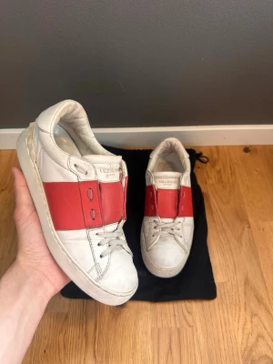 Valentino vita sneakers med röd detalj - Säljer ett par vita Valentino sneakers i läder med bred röd rem över mitten och klassisk logga på plösen. Skorna har lågt skaft och vita snören. Snygg och stilren design som sticker ut med den röda detaljen. Perfekta för dig som gillar exklusiva märken och streetstyle.