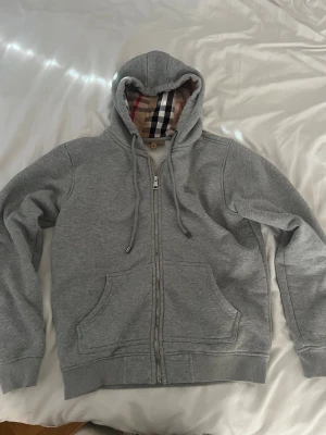 Burberry ziphoodie - Säljer en grå Burberry hoodie med dragkedja framtill, två stora fickor och broderad logga på bröstet. Hoodien har justerbar huva med snören och ribbade muddar vid ärmar och nederkant. Tillverkad i mjuk bomullsblandning för extra komfort.