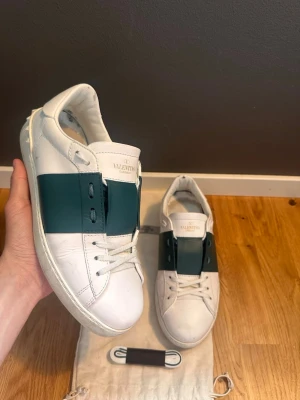Valentino vita sneakers med grön detalj - Snygga vita sneakers från Valentino med en bred grön rem över mitten. Skorna är låga och har klassisk snörning samt diskret logga på plösen. Tillverkade i läder och kommer med extra skosnören och dustbag. Perfekta för dig som vill ha en clean men unik look.