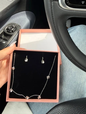 Blomformat halsband från Edblad - Säljer ett silvrigt halsband från Edblad med små blommor som dekoration. Halsbandet har en tunn kedja och blommorna har en glittrande detalj i mitten. Kommer i original ask tillsammans med matchande örhängen. Perfekt för dig som gillar stilrena och söta smycken.
