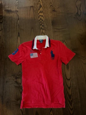 Ralph Lauren polo  - Röd pikétröja med klassisk krage i vitt och blått från Polo Ralph Lauren. Tröjan har korta ärmar, broderad logga på bröstet, amerikansk flagga och detaljer i blått. Perfekt för en sportig och stilren look. Passar M ochså 