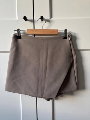 Asymmetrisk byxkjol från Zara - Stilren grå skort med omlott-detalj framtill och diskreta fickor bak. Skorten har en rak passform och stängs med dragkedja i sidan. Tillverkad i ett mjukt, vävt material som ger en clean och modern look. Använt en gång💞
