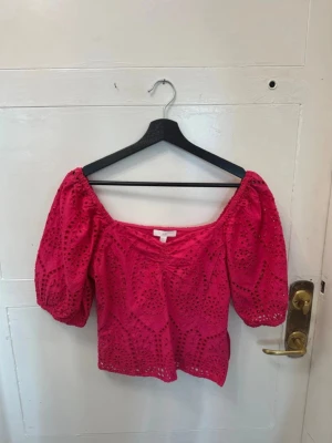 Rosa broderad blus med puffärm - Trendig rosa blus med broderade detaljer och puffiga korta ärmar. Toppen har en v-ringad urringning och är tillverkad i ett lätt material som passar perfekt till sommaren. Snygg att styla med både jeans och kjol.
