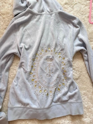 Ljusblå Juicy Couture hoodie - Säljer en ljusblå hoodie från Juicy Couture i mjuk velour. Dragkedja framtill, kängurufickor och huva. Baksidan har ett stort glittrigt emblem med stenar och Juicy Couture-logga. Perfekt för dig som gillar en lyxig och chill stil. Fläck