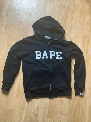 Svart BAPE zip hoodie med camo-apa - Säljer en svart hoodie från BAPE med dragkedja, stor vit BAPE-logga på bröstet och ikoniskt camo-apansikte på ryggen. Hoodien har huva, muddar vid ärmslut och nederkant samt en stor magficka. Perfekt för dig som gillar streetwear och vill sticka ut. A bathing APE
