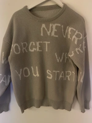 Grå stickad tröja med text - Cool grå stickad tröja med vit text som säger 'NEVER FORGET WHY YOU START'. Tröjan har rund halsringning, långa ärmar och ribbstickade muddar vid ärmslut och nederkant. Perfekt för en avslappnad och trendig look.