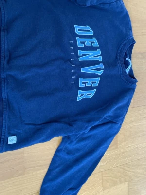 Mörkblå sweatshirt med Denver-tryck - Mörkblå sweatshirt från 157 med stort tryck 'DENVER COLORADO' i ljusblått och vitt på bröstet. Tröjan har rund halsringning, ribbade muddar vid ärmslut och nederkant samt mjukt material på insidan. Perfekt för en avslappnad och sportig stil.